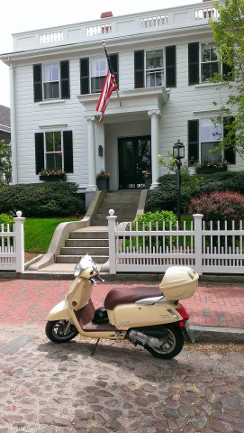 A 2 story white colonial building with concrete stairs leading down to a white picket fence. A yellow scooter is parked on the cobblestone street