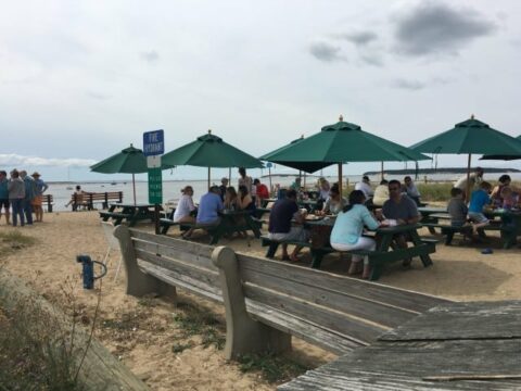 an outdoor restaurant by the beach with wooden picnic tables topped with green umbrellas and people eating
